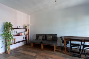 A living room with a sofa, a wooden table, and a bookshelf with plants and books. - Casa com Churrasqueira a 550m da Praia Itaipua u (Marica)