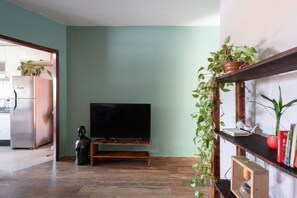 Uma sala de estar com uma televisão sobre um suporte de madeira, uma cozinha visível por uma porta aberta e uma estante com plantas na parede.
