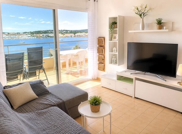 A modern living room with a large TV, a sofa, a small table with a plant, and a balcony with a view of the sea. - Sea View Apartment - Swimming pool and tennis (L'Escala)