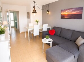 A modern living room with a grey sofa, a dining table with chairs, a small white table with a plant and remote, and a sunset painting on the wall. - Sea View Apartment - Swimming pool and tennis (L'Escala)