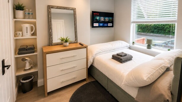 Un dormitorio con cama, cómoda, espejo, televisor mostrando Netflix y ventana con persianas.