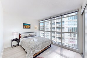A bedroom with a large window, a bed with a patterned bedspread, a nightstand with a lamp, and a painting on the wall. - Over 1000 SF Condo, free parking (Toronto)