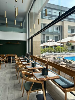 A modern cafe with wooden furniture, a pool view, and hanging pendant lights. - R&P Suites @ Embassy Gardens (Accra)