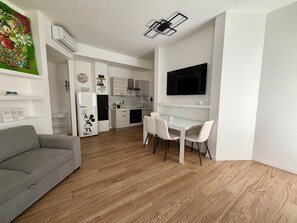 Ein modernes Wohnzimmer mit grauem Sofa, einem Essbereich mit weißen Stühlen, einer Küche und Holzboden.