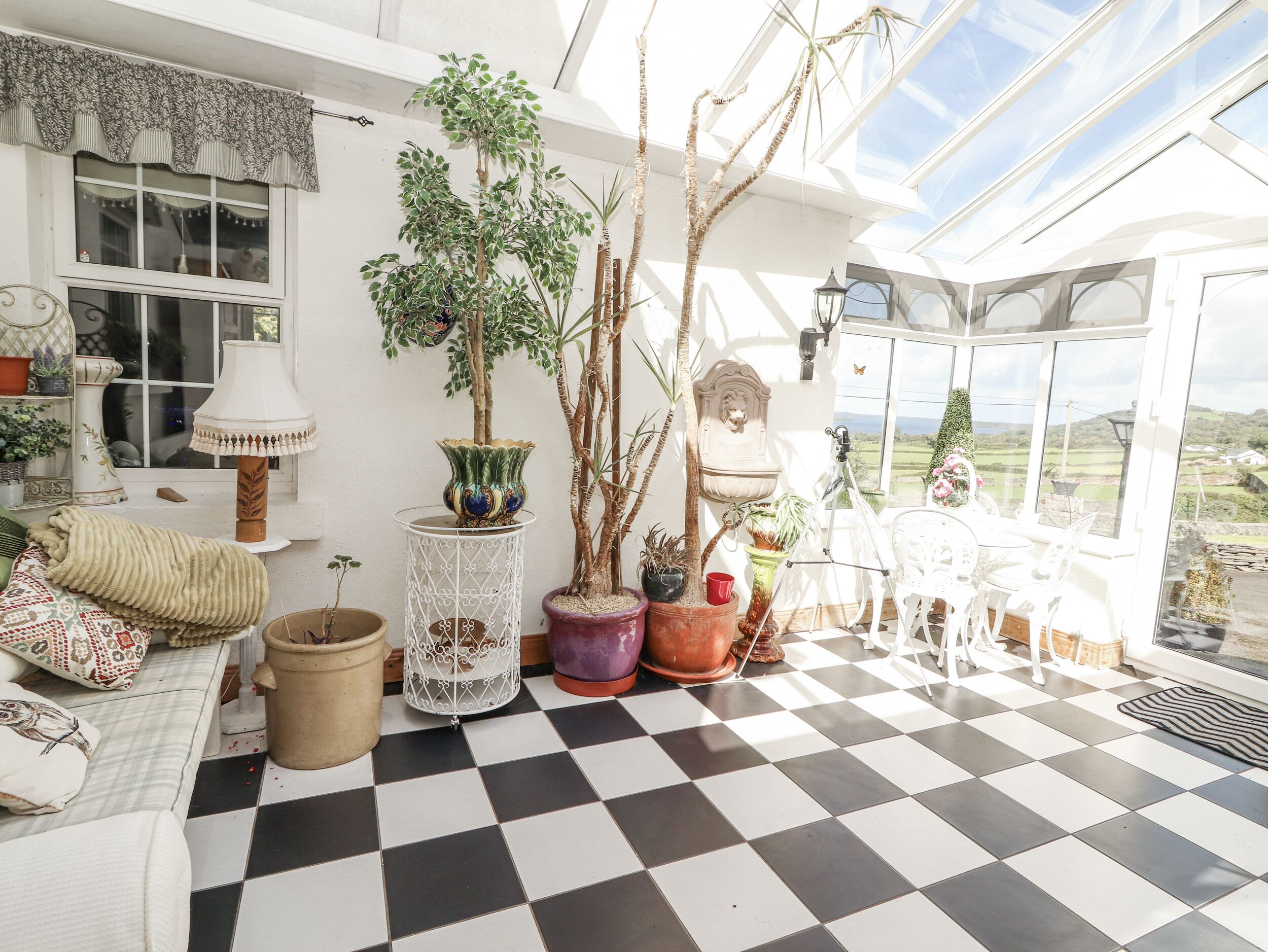 A conservatory with a checkered floor, potted plants, a white sofa, and a view of the countryside.