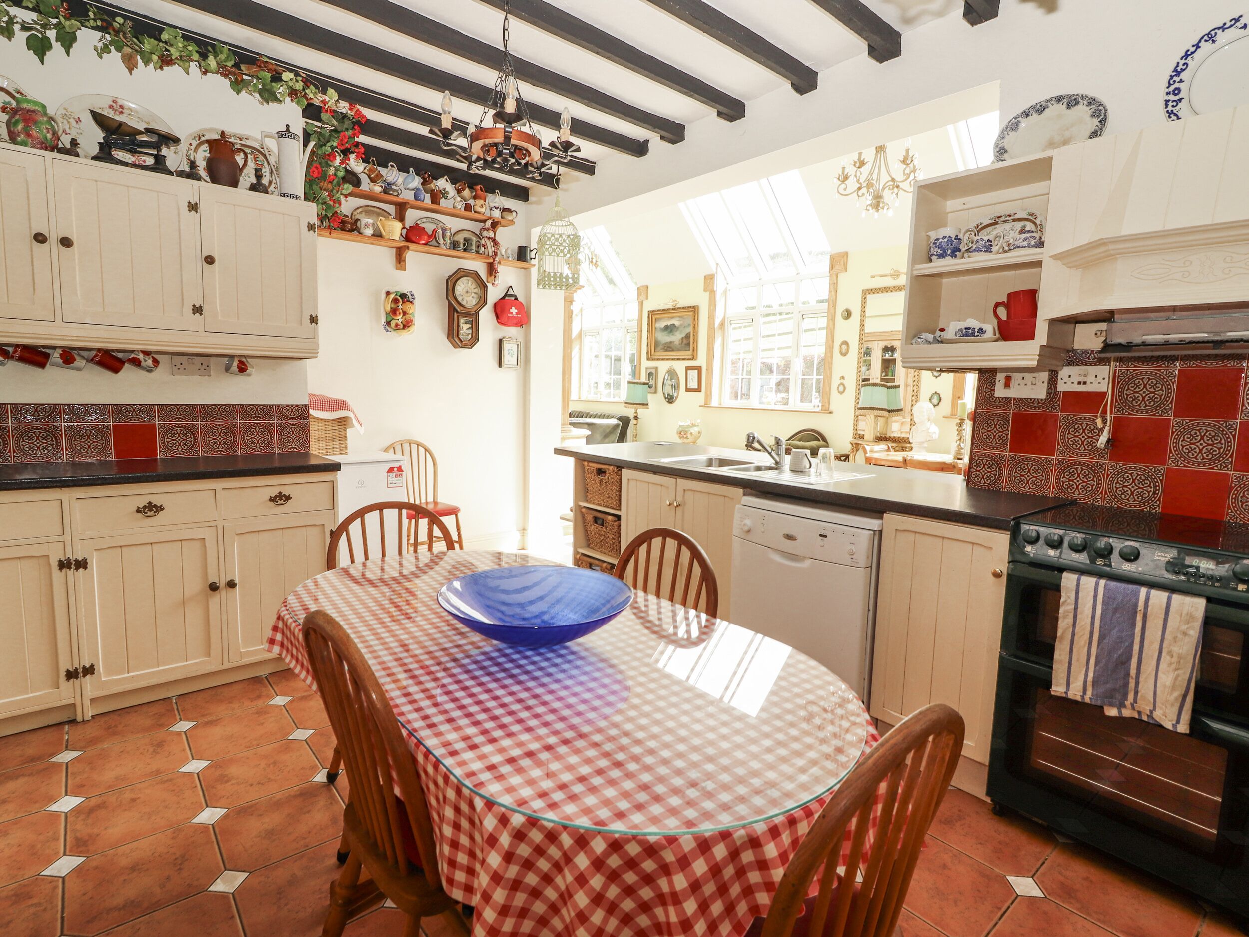 A kitchen with a checkered tablecloth, a blue bowl, wooden chairs, a stove, and various kitchen items on shelves.