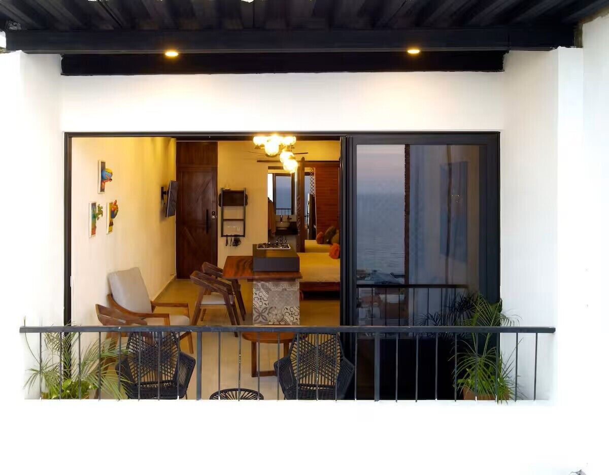 Un balcón con zona de comedor, un sofá y una cocina visible a través de un ventanal corredero.