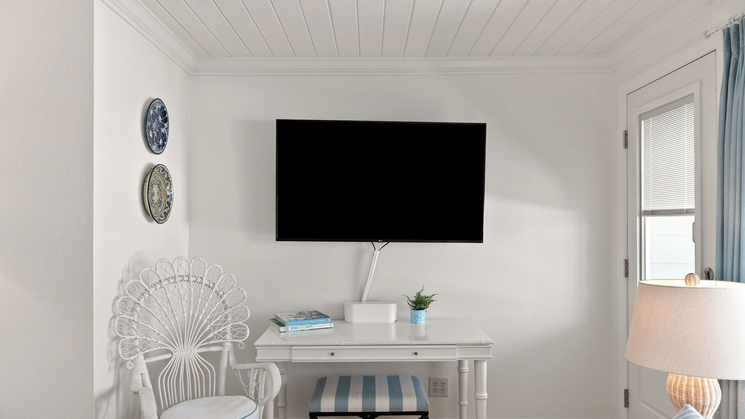 Ein Zimmer mit weißem Schreibtisch, einem Sessel, einem an der Wand montierten Fernseher und einem Fenster mit Vorhängen.