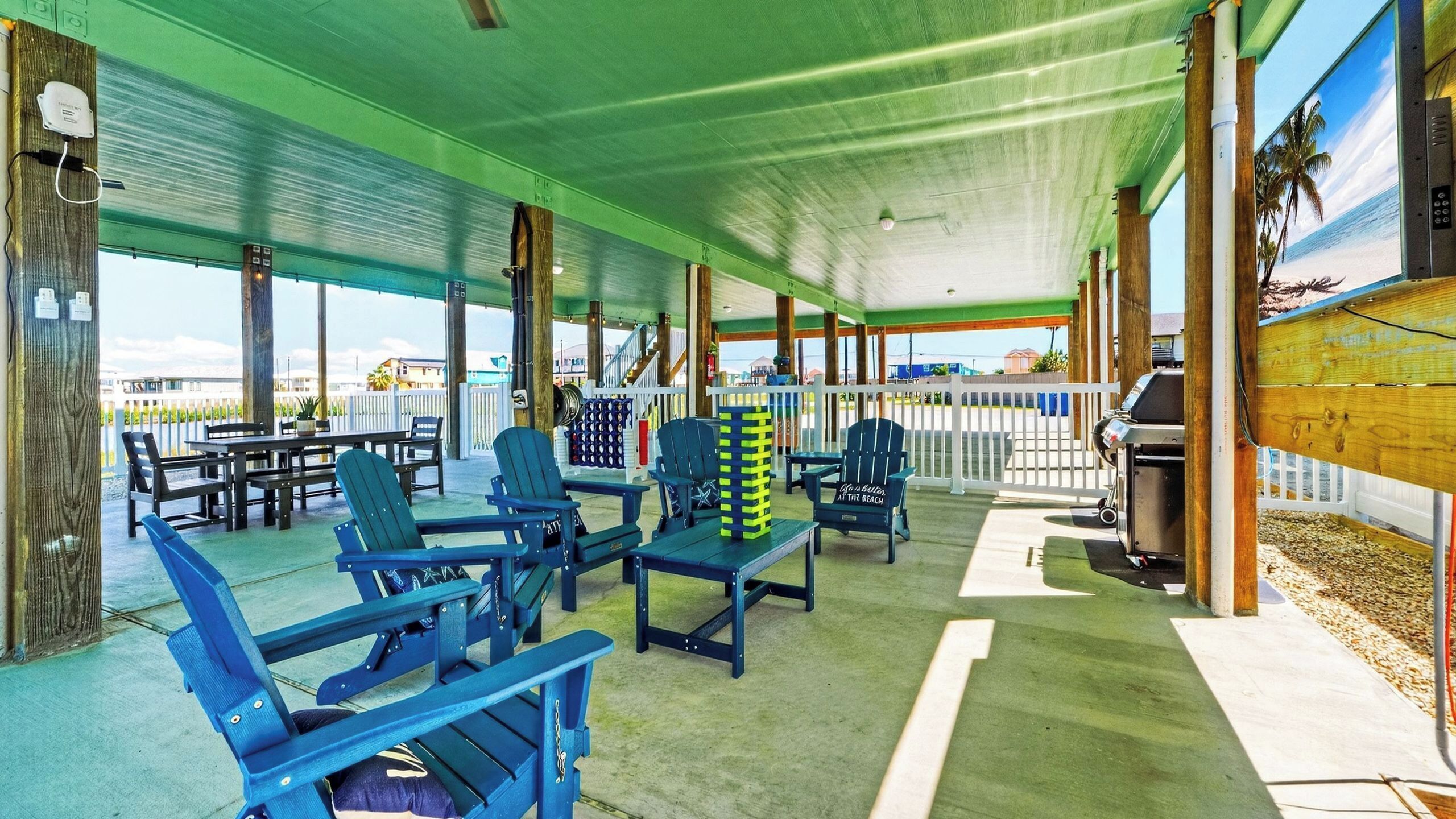 A covered outdoor seating area with blue Adirondack chairs, a barbecue grill, and a view of the beach.