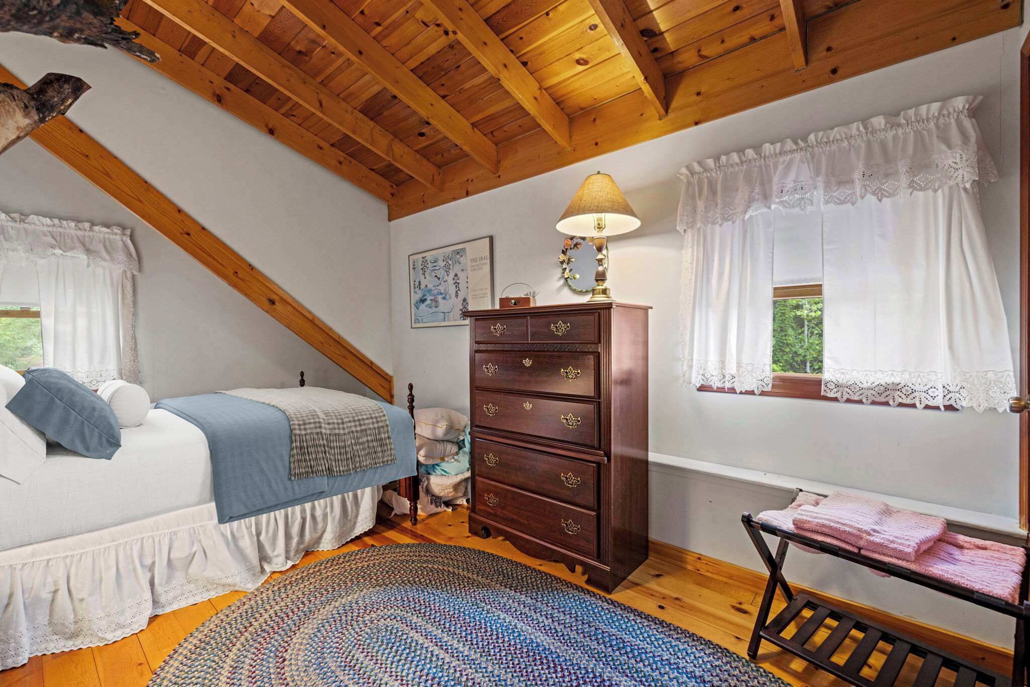 A bedroom with a wooden ceiling, a bed with a blue blanket, a dresser, a lamp, and a window with lace curtains.
