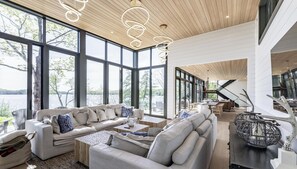 Een ruime woonkamer met een groot raam met uitzicht op een meer, een moderne kroonluchter en een eethoek met een trap.