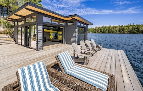 Una moderna casa flotante con terraza, sillones de descanso y una zona cubierta pequeña.
