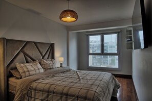 Ein Schlafzimmer mit einem großen Bett, einem Kopfteil, einem Nachttisch mit Lampe, einem Fenster mit Blick auf die Stadt und einem an der Wand montierten Fernseher.