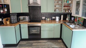 A kitchen with teal cabinets, a black backsplash, a stainless steel range hood, a microwave, a toaster, a sink, and a window. - Four Seasons (Grimsby)