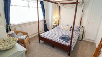 A four-poster bed with a striped bedspread, a wicker chair, a table with a bowl, and a radiator.