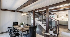 Un espace repas avec une table en bois, des chaises noires et un lustre. Un escalier avec une rampe en bois mène à la cuisine.