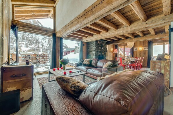 Ein gemütliches, rustikales Interieur mit Steinwand, Holzbalken, Ledermöbeln und Blick auf eine verschneite Landschaft.