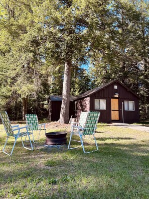 A rustic cabin with a large tree in front, two checkered chairs, and a fire pit. - Birch at Hiawatha Cabins (Munising Township)
