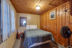 Une pièce aux murs lambrissés, comprenant un lit, une petite table de chevet, un miroir, un tableau accroché au mur et un ventilateur sur pied.