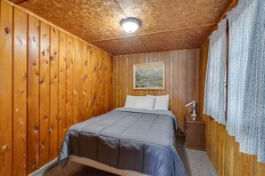 A wooden-paneled room with a bed, a nightstand, a lamp, and a painting on the wall. - Birch at Hiawatha Cabins (Munising Township)