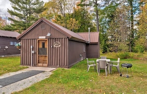 A cabin with a door labeled 'HEIMLOCK,' a small outdoor seating area with a table and chairs, and a grill.
