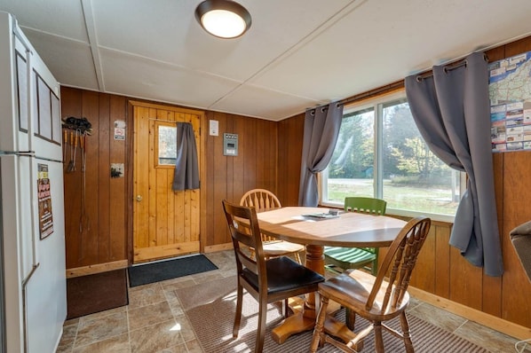 A cabin-style room with a wooden interior, a dining table with chairs, a refrigerator, a window with curtains, and a map on the wall.