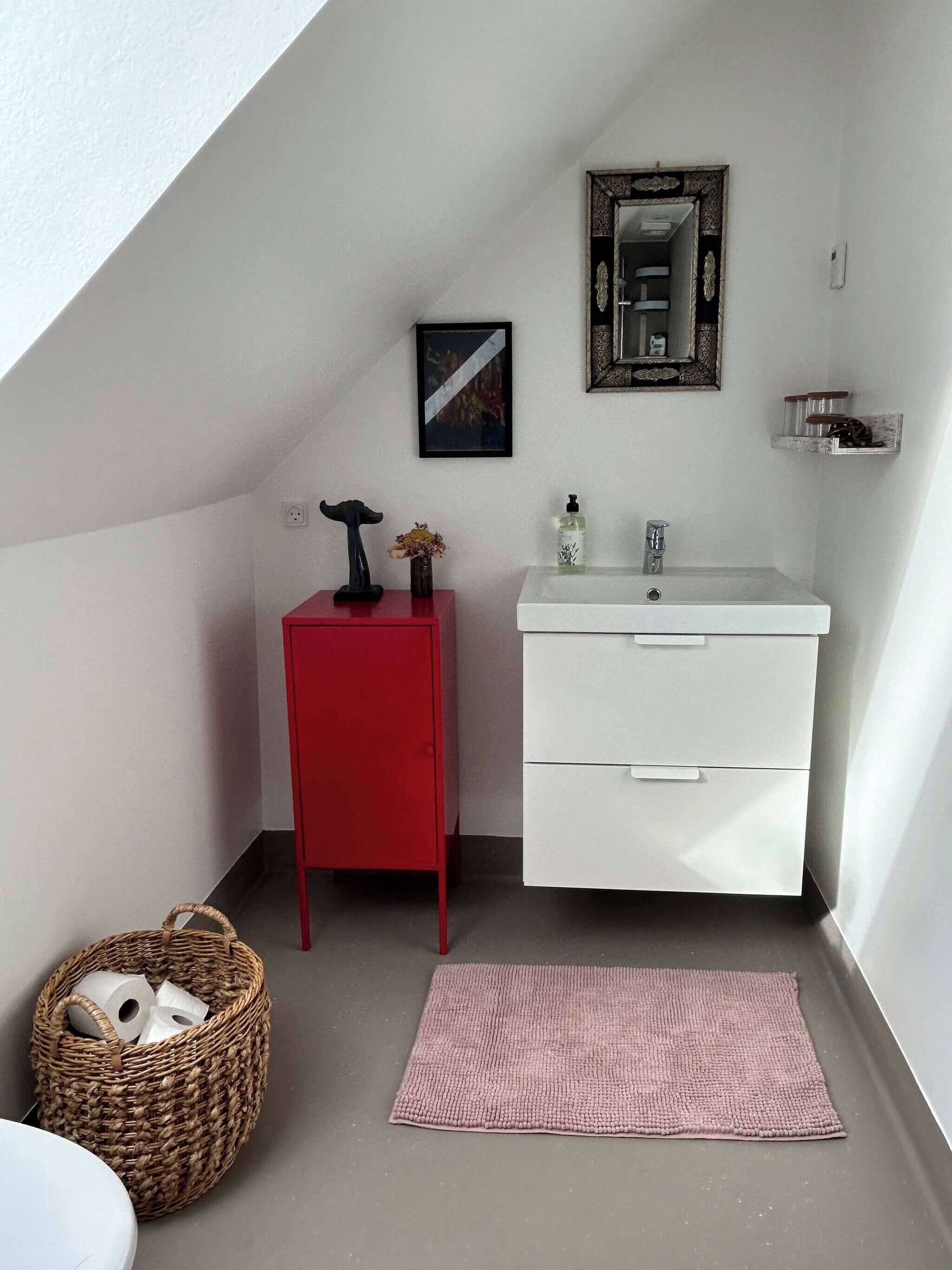 Un baño pequeño con un lavabo blanco, un mueble rojo y un espejo encima. Hay una cesta de mimbre en el suelo y una alfombra rosa.