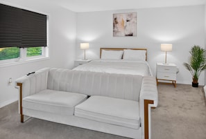 Une chambre à coucher comprenant un lit, des tables de chevet, un canapé, une fenêtre avec un store, une œuvre d’art murale et une plante en pot.