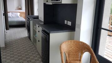 A compact kitchen with a dining area, a wicker chair, and a bed in the background.