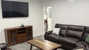 Ein Wohnzimmer mit braunem Ledersofa, einem Holztisch, einem Ledersessel und einer Holzkommode. Ein Flachbildfernseher ist an der Wand befestigt.