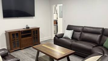 Un salon avec un canapé en cuir marron, une table basse en bois, un fauteuil en cuir et un meuble en bois. Un téléviseur à écran plat est fixé au mur.