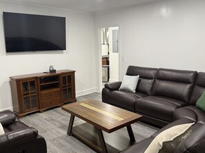 Ein Wohnzimmer mit braunem Ledersofa, einem Holztisch, einem Ledersessel und einer Holzkommode. Ein Flachbildfernseher ist an der Wand befestigt.
