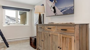 A room with a large window, a wooden cabinet, a lamp, and a framed picture of a skier. - Elegant Avon Center Condo I Walk to gondola (Avon)