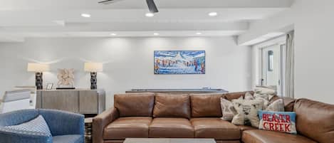 A living room with a brown leather sofa, a blue armchair, a coffee table with magazines, a ceiling fan, recessed lighting, and a painting on the wall.