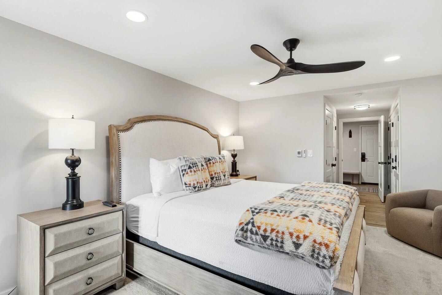 A bedroom with a bed, bedside tables, lamps, a ceiling fan, and a door leading to another room.