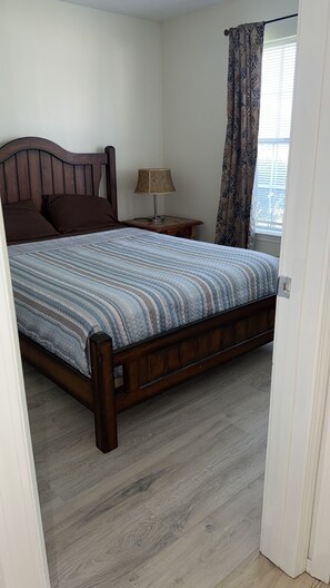 A bedroom with a wooden bed, a bedside table with a lamp, a window with curtains, and a door.