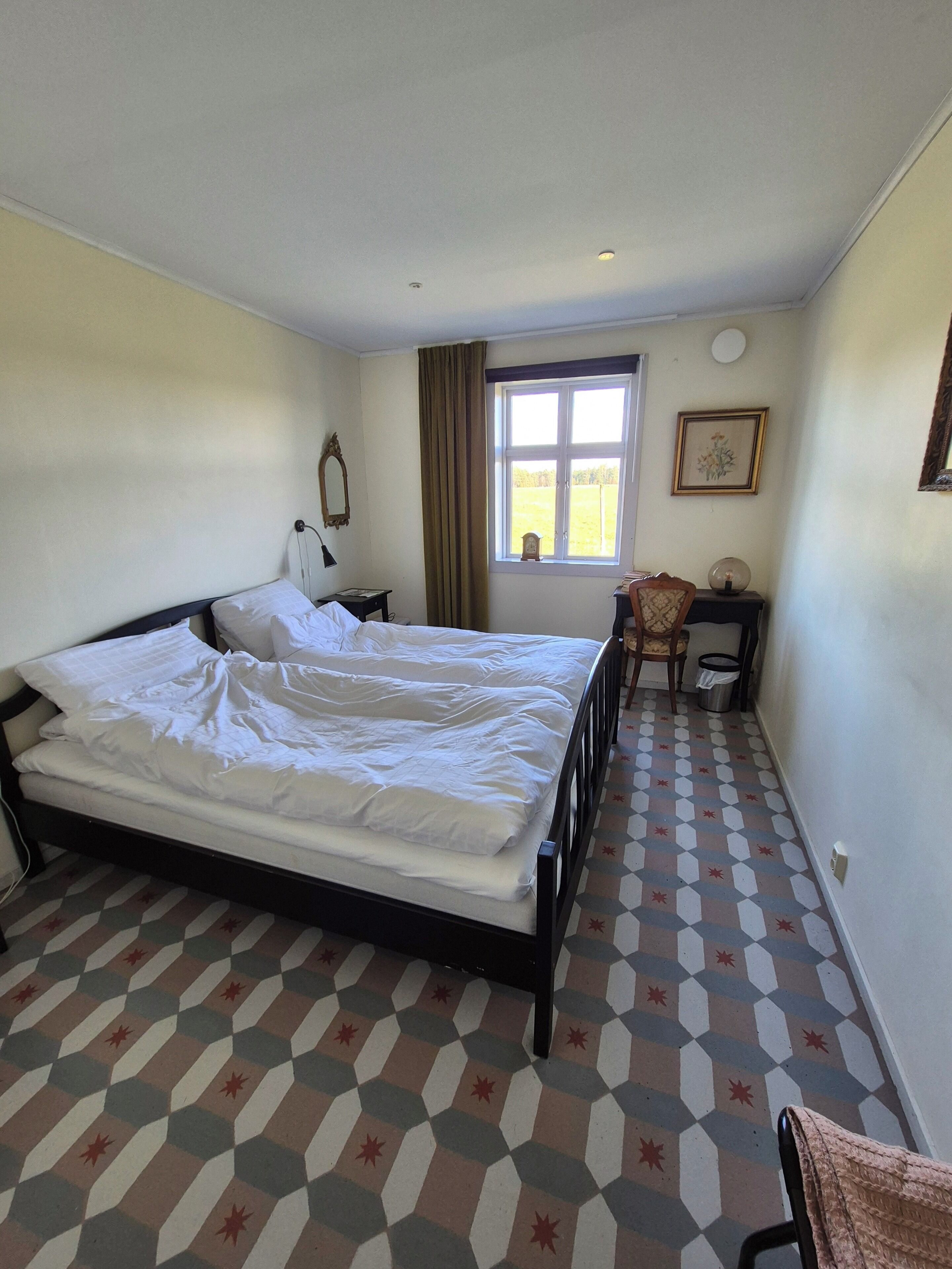 Un dormitorio con una cama, una ventana, una silla, un escritorio y suelo estampado.