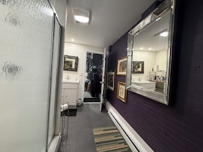 Un pasillo estrecho con una pared morada oscura, una puerta blanca, un espejo, un lavabo y un pequeño cubo de basura.