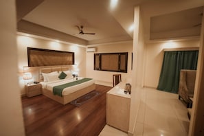 A bedroom with a bed, bedside tables, a desk, a chair, a ceiling fan, and a window with curtains. - D'polo Club & Spa Resort (Dharamshala)