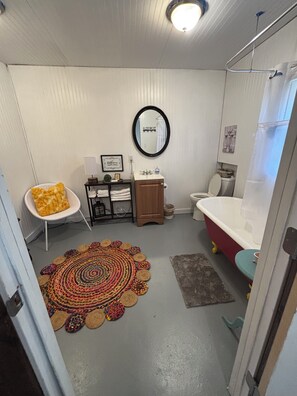 Une petite salle de bain avec un meuble-lavabo blanc, un miroir rond, une baignoire rouge et un tapis coloré.