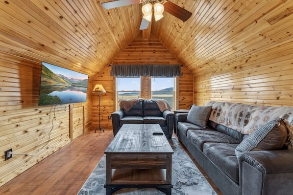 Interior de una cabaña de troncos con techo de madera, un ventanal grande, un sofá, una mesa de centro y un televisor.
