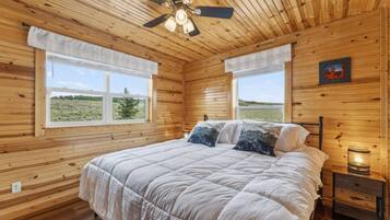 A wooden cabin bedroom with a bed, two windows, a ceiling fan, and a nightstand.