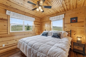 A wooden cabin bedroom with a bed, two windows, a ceiling fan, and a nightstand. - Elk Ridge Retreat | Hot Tub & 360° Views (Park County)