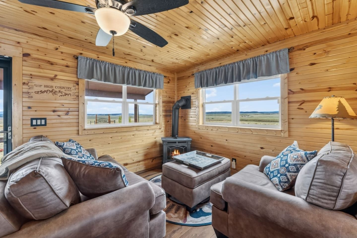 L’intérieur d’un chalet accueillant, avec des murs en bois, deux fenêtres avec des rideaux, un ventilateur de plafond, une cheminée et deux canapés ornés de coussins à motifs.