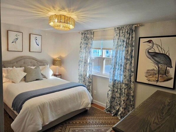 Quarto com cama, lustre, janela com cortinas e obras de arte emolduradas nas paredes.