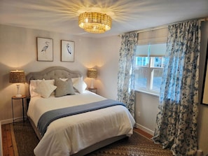 Une chambre Ă coucher comprenant un lit, deux lampes fixĂ©es au mur, un lustre, une fenĂȘtre avec des rideaux et deux tableaux reprĂ©sentant des oiseaux, Ă©galement fixĂ©s au mur.