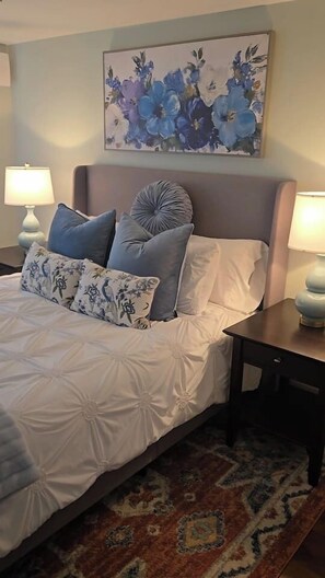 Un dormitorio con cama, almohadas, mesita de noche, lámpara, un cuadro floral y una alfombra estampada.