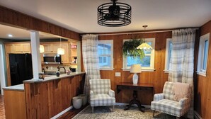 A cozy living room with a wooden floor, a central rug, two plaid chairs, a small table with a lamp, and a kitchen area visible through an open doorway. - The Heron's Perch - HL (Hampton)