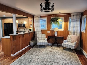 A kitchen with wooden cabinets, a black refrigerator, and a microwave. There are two plaid chairs and a small table with a lamp.