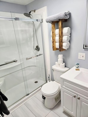 A bathroom with a glass-enclosed shower, a toilet, a sink, and a towel rack with folded towels. - The Heron's Perch - HL (Hampton)